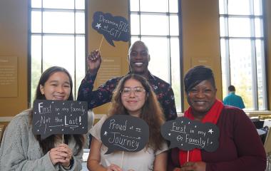 Associate Director of First-Generation Student Initiatives Charmaine Troy (far right) holds up sign that reads "First in my family" with students and MC from First-Generation Symposium.